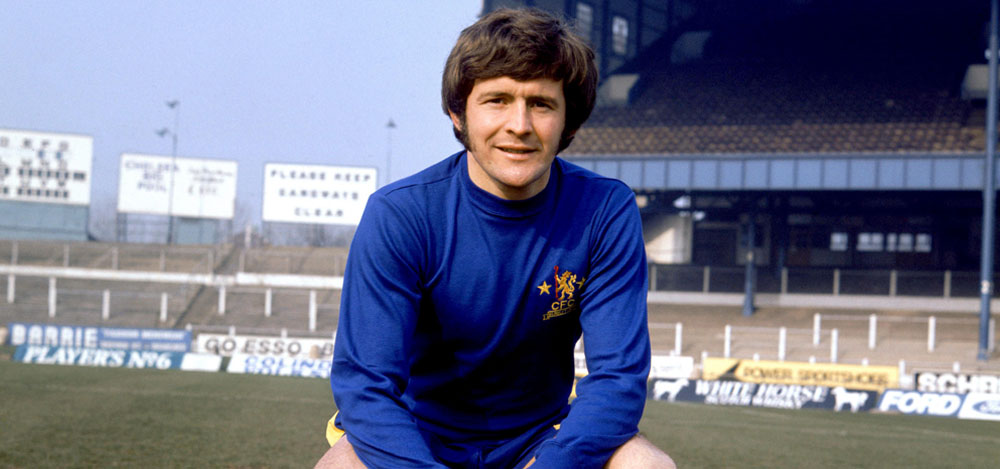 John Hollins in a blue Chelsea top sits on a football pitch in a football ground. 